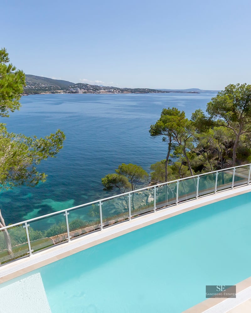 Rectangular turquoise swimming pool overlooking the Mediterranean sea with pine trees and a glass railing.