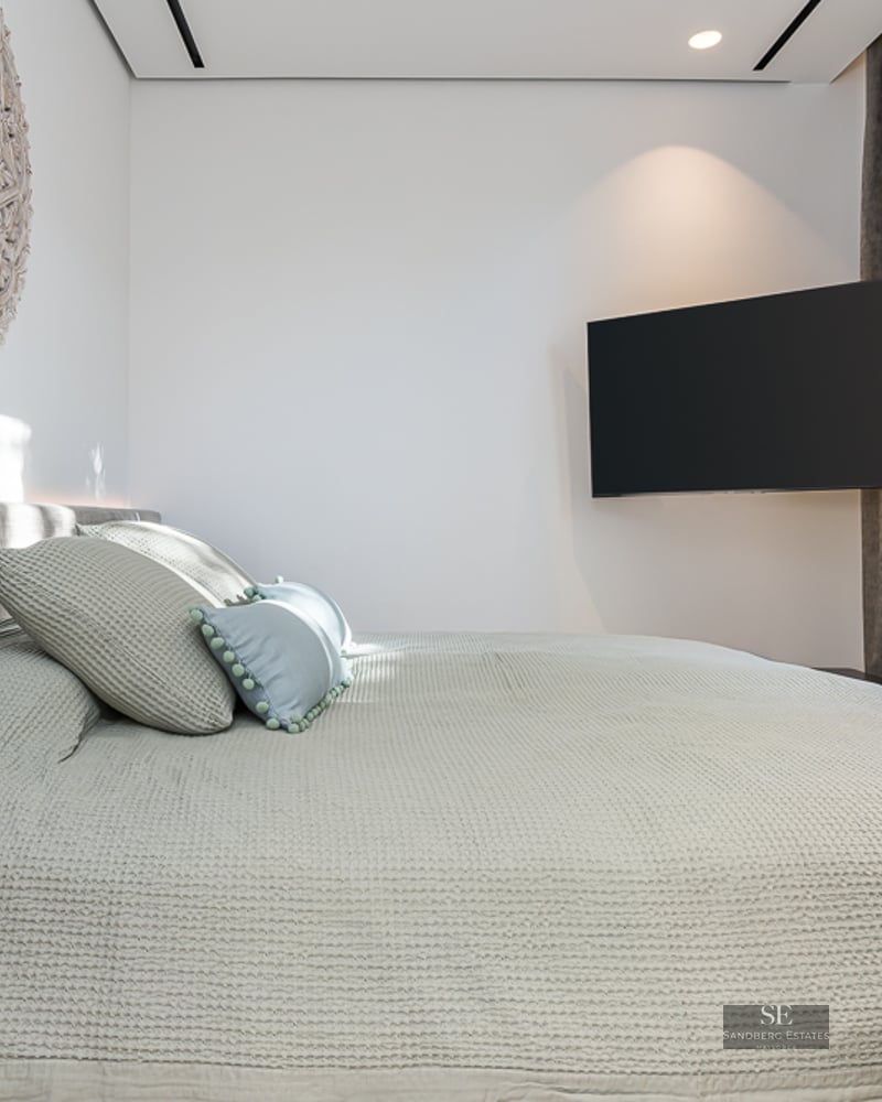 Modern bedroom featuring a grey bed, wall-mounted TV, and a large circular carved wooden ornament on a white wall.