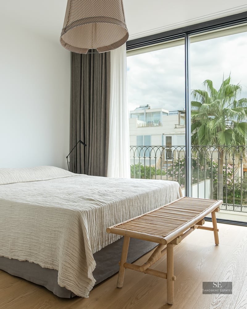 Bright bedroom with floor-to-ceiling windows, light wood floors, a large bed, and a palm tree view outside.
