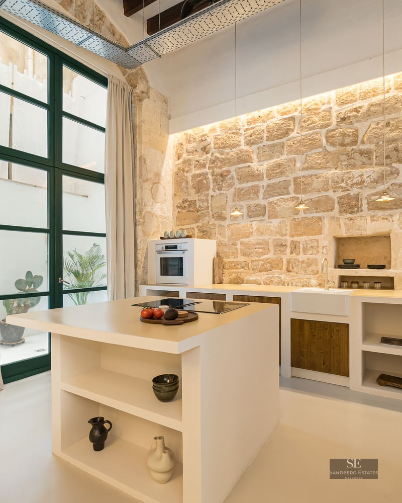 Modern kitchen with a central island set against a large rustic exposed stone wall and tall windows.