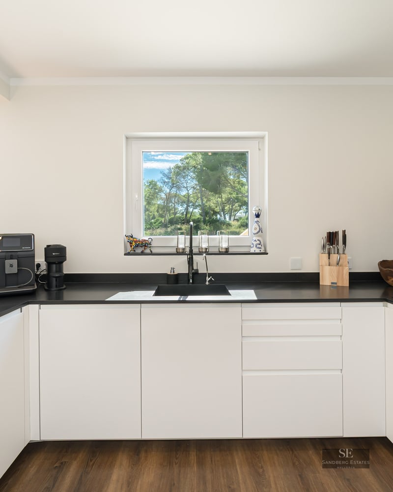 Cuisine moderne avec placards blancs, plans de travail noirs et une fenêtre donnant sur des arbres verts.
