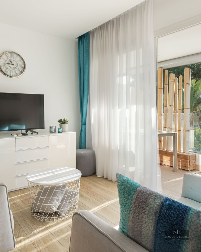 Salon moderne avec canapés gris, coussins turquoise et grandes baies vitrées ouvrant sur une terrasse arborée.