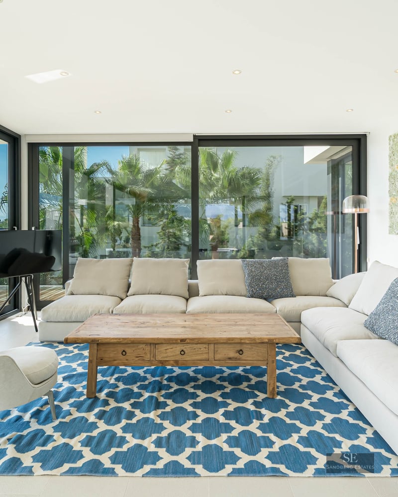 Modern living room with white L-shaped sofa, blue patterned rug, and floor-to-ceiling windows showing a coastal view.