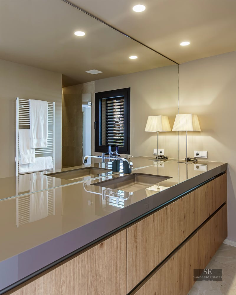 A large mirror reflects a modern wooden bathroom vanity with an integrated sink and designer table lamps.