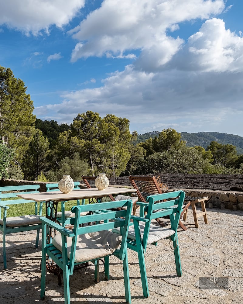 Stone terrace with a white table and turquoise chairs surrounded by forest and mountains under a blue sky.