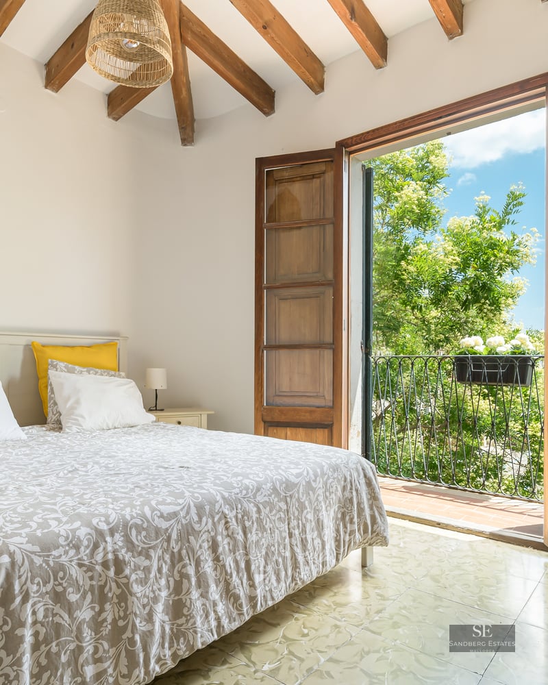 A bright bedroom with exposed wooden ceiling beams and French doors opening to a sunny balcony with green trees.