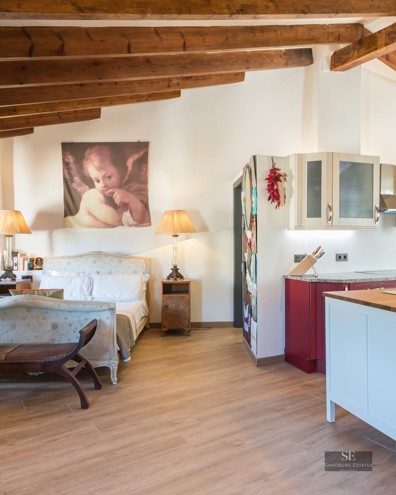 Studio featuring a classic bed, exposed wooden ceiling beams, a stone wall, and a modern red and white kitchen with island.