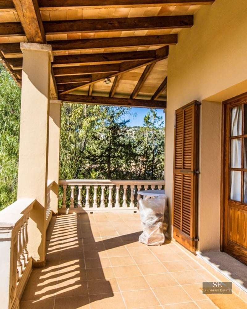 Covered terrace with wooden beams, tiled floor, and stone balustrade overlooking a lush green landscape.