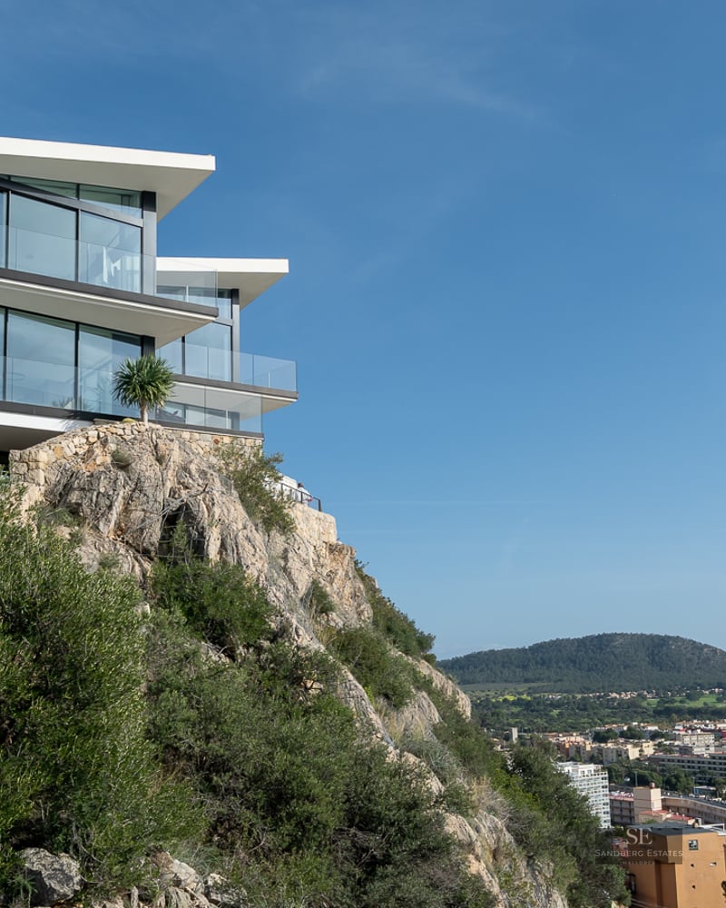 Modern white villa with large glass windows perched on a rocky cliff overlooking a city landscape under a blue sky.