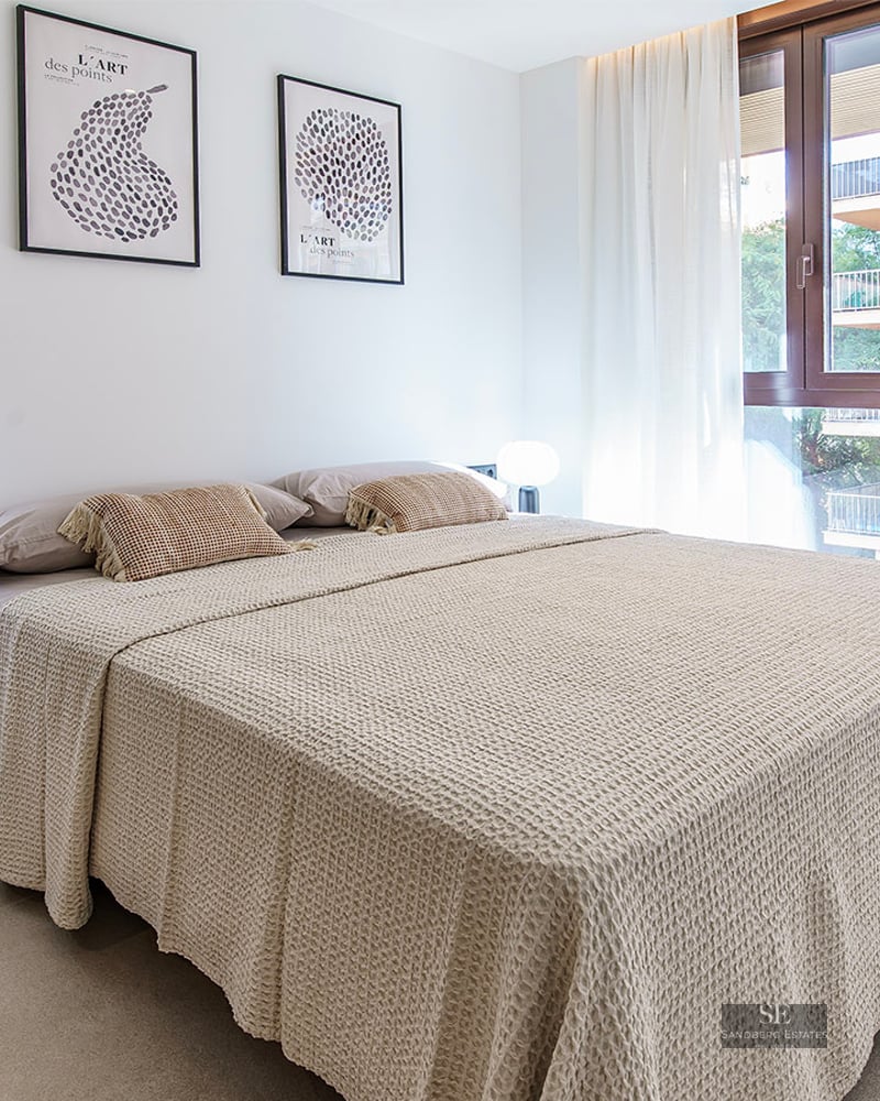 Bright bedroom with a large bed, beige waffle-knit linens, designer lamps, and a window with sheer white curtains.