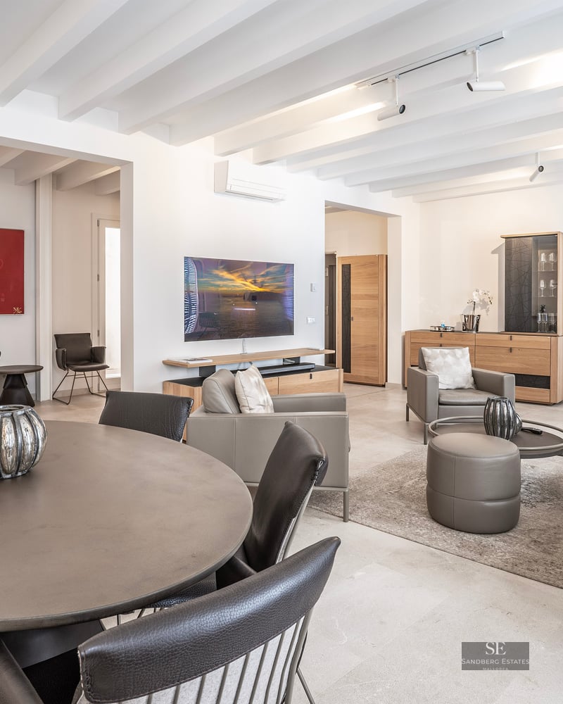 Modern open-plan living area with a round dining table, leather sofas, and white beamed ceiling.
