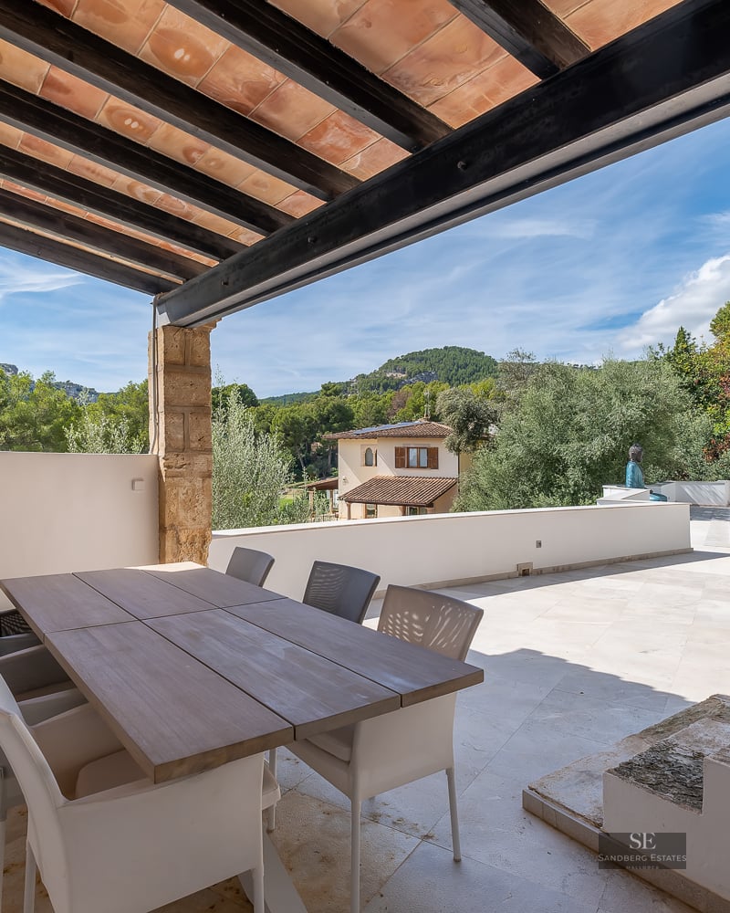 Outdoor dining area under a wooden beam pergola overlooking a pool, garden, and distant hills.
