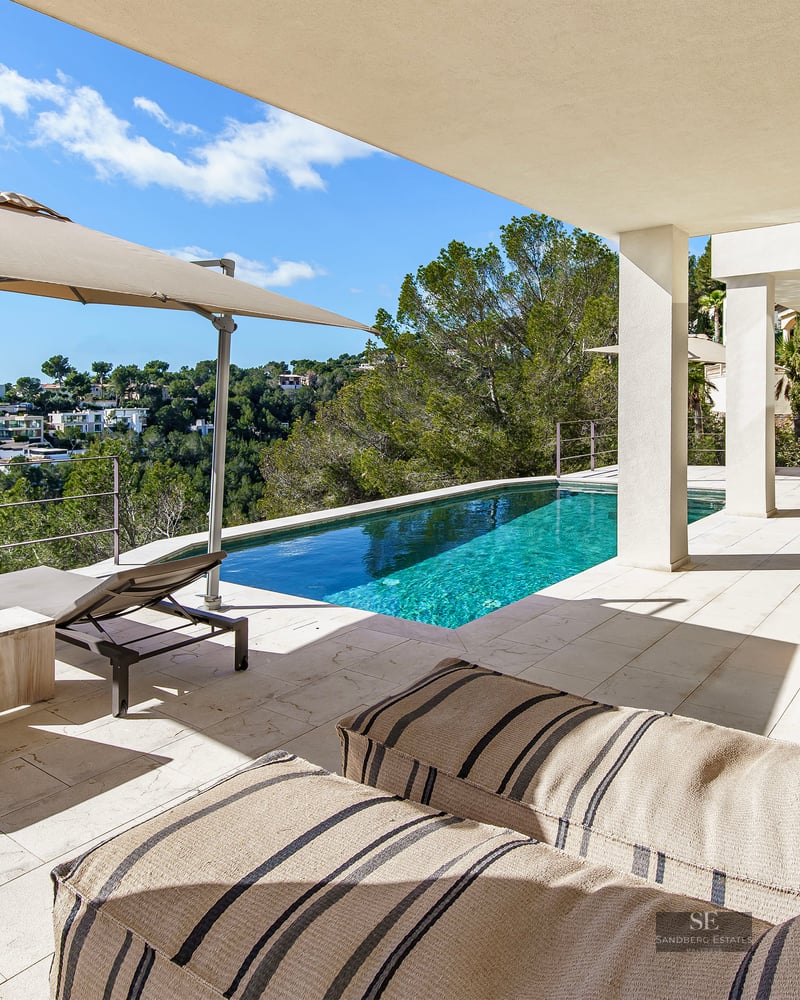 Modern swimming pool on a stone terrace with sun loungers and a large umbrella overlooking a green hillside.