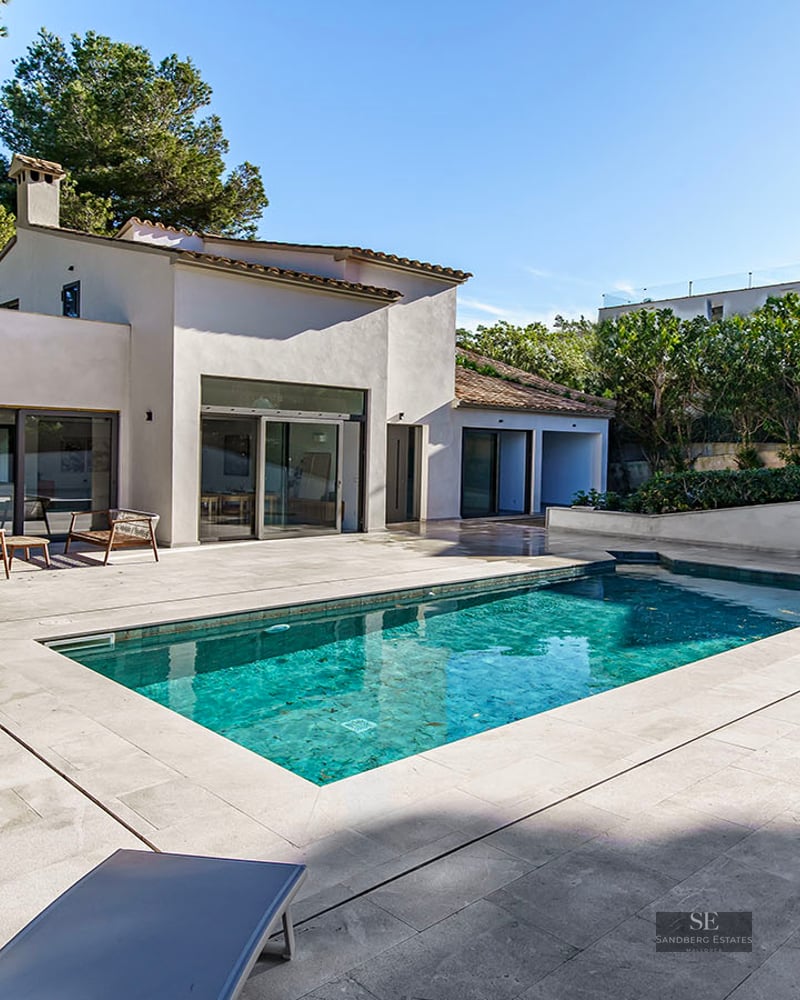 Modern white villa with a turquoise rectangular swimming pool and stone terrace surrounded by pine trees.