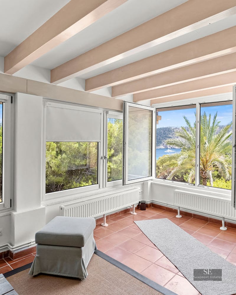 Interior room with terracotta floor tiles, wooden ceiling beams, and large windows overlooking pine trees and the sea.