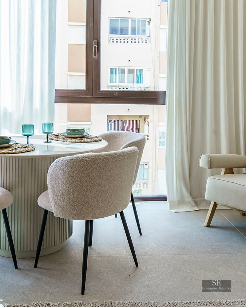 Bright dining room with round white table, four bouclé chairs, large windows with white curtains, and a beige armchair.