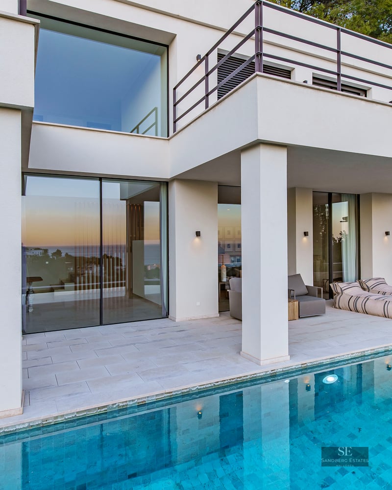 Modern white villa with a turquoise swimming pool and large glass windows reflecting a sunset.