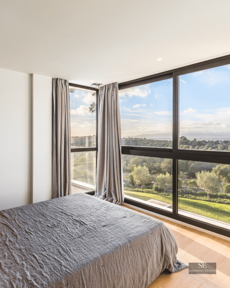 Modern bedroom with large bed and floor-to-ceiling windows overlooking a green landscape and blue sky.