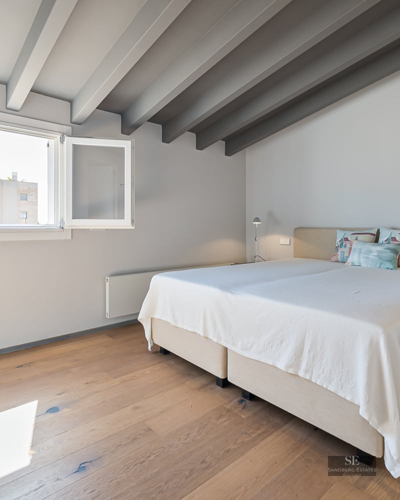 Bright bedroom featuring a double bed, wooden flooring, grey ceiling beams, and a large window.