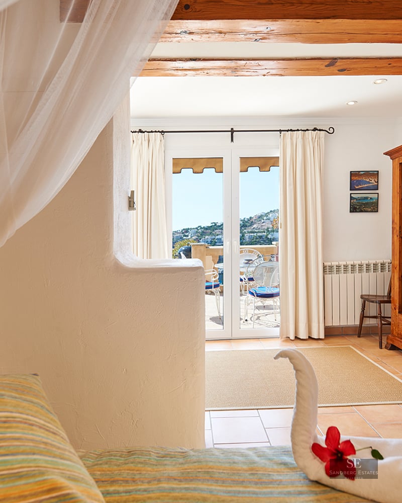 A rustic bedroom with exposed wood beams, a canopy bed with a towel swan, and doors opening to a sunny terrace.