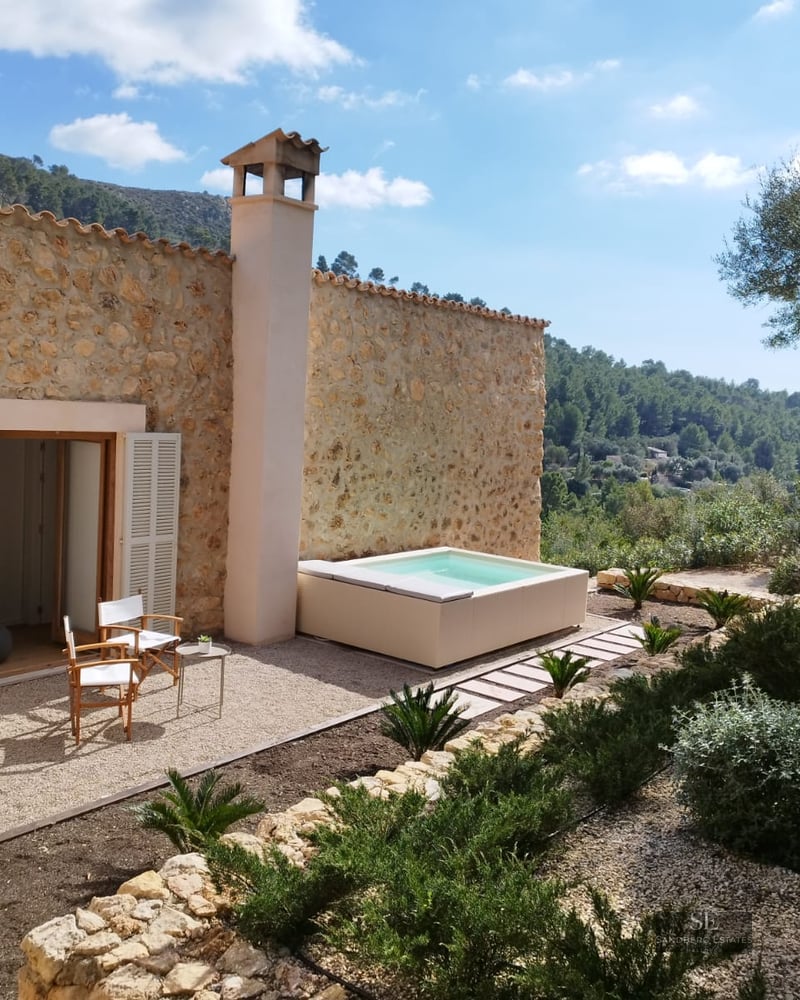 Stone house featuring an outdoor jacuzzi, wooden chairs on gravel, and mountain views under a clear sky.