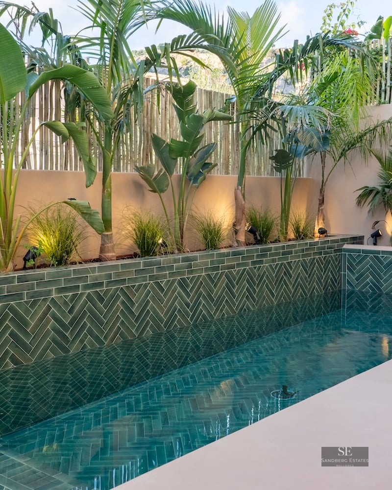 Piscine de luxe avec carreaux verts en chevrons, palmiers et murs beiges avec un éclairage chaleureux.