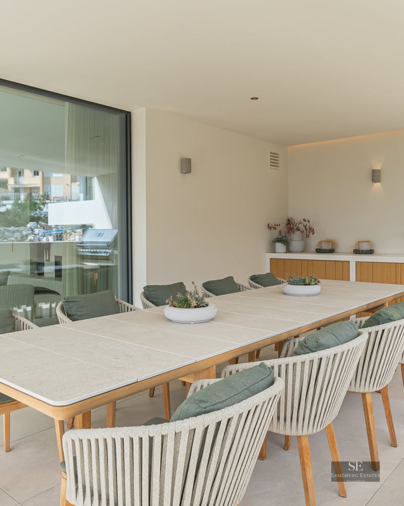 Large outdoor dining table with ten chairs next to an outdoor kitchen and grill on a modern covered terrace.