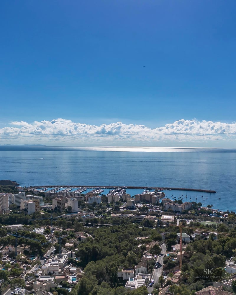 Vue aérienne d'une ville côtière avec marina, arbres verts et mer Méditerranée sous un ciel bleu clair.