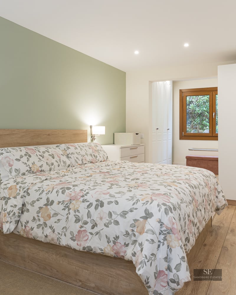 Modern bedroom featuring a floral bed, sage green wall, white furniture, and light wood flooring.