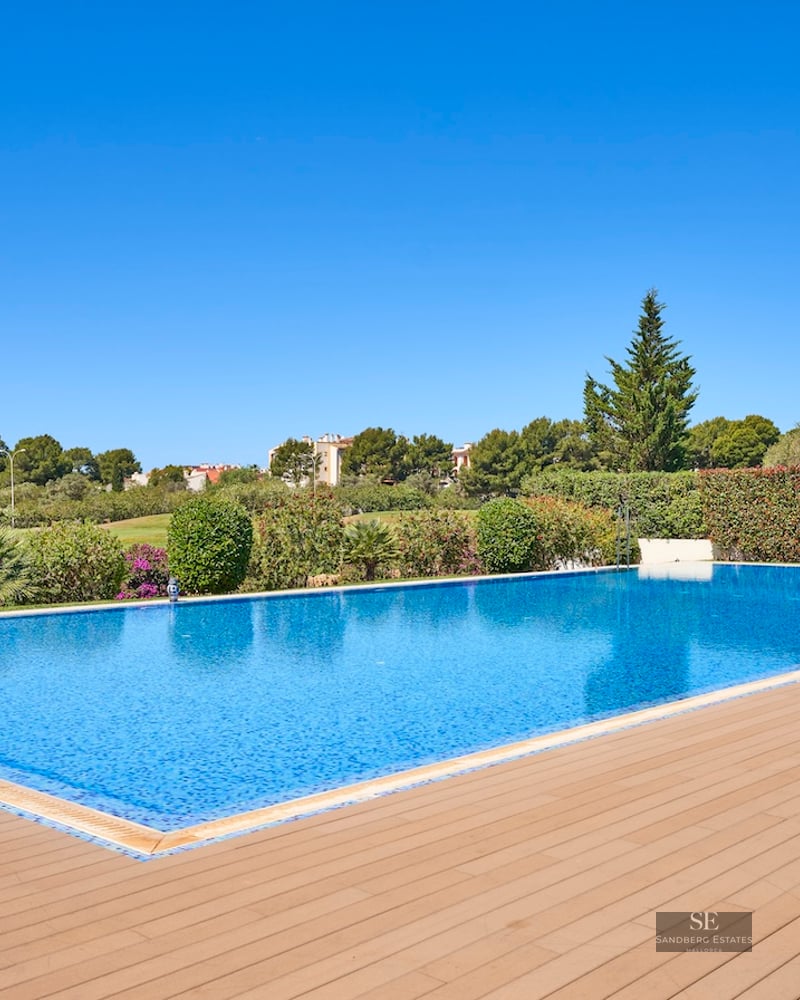 Large blue swimming pool with a wooden deck, surrounded by green hedges and a Mediterranean villa under a clear sky.