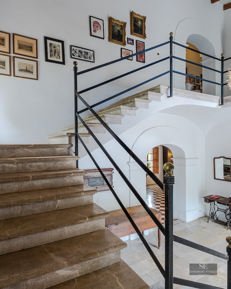 Grand entryway featuring a stone staircase, white walls adorned with art, and a mezzanine library level.