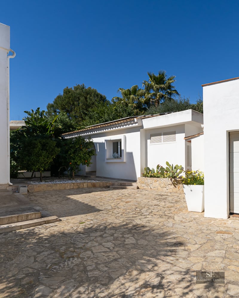White Mediterranean villa with a stone-paved driveway, garage, and lush greenery under a clear blue sky.