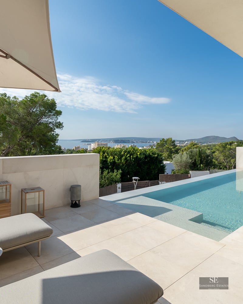 Sun loungers on a modern beige stone terrace next to a swimming pool overlooking the sea and green landscape under a clear sky.