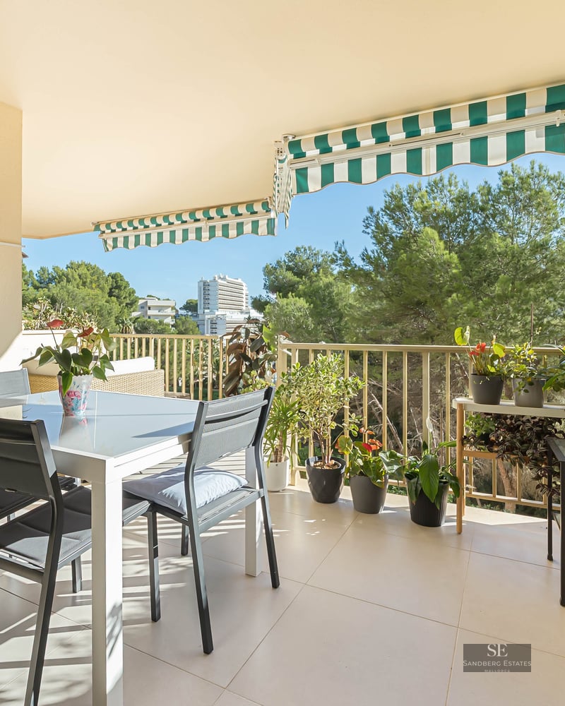 Un balcon ensoleillé avec une table à manger blanche, des chaises sombres, de nombreuses plantes en pot et des stores rayés.