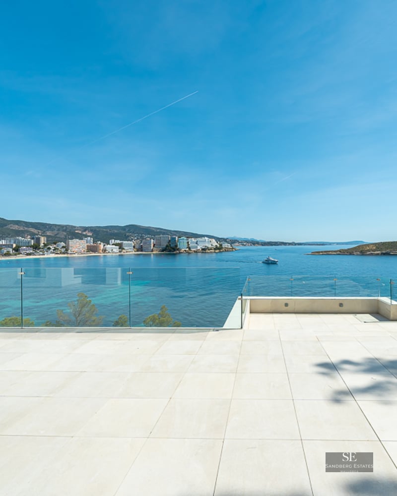 Large white tiled terrace with glass railings overlooking a blue bay, mountains, and coastal town.