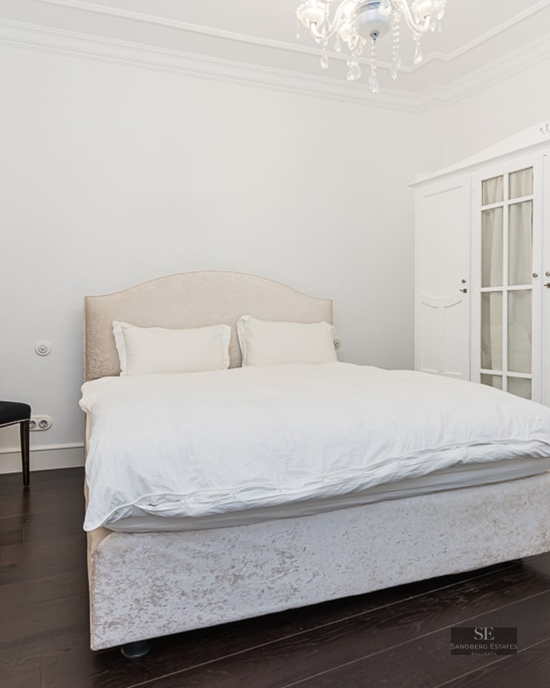 Bright bedroom featuring a beige bed, large white wardrobe, black chair, and crystal chandelier on dark wood floors.