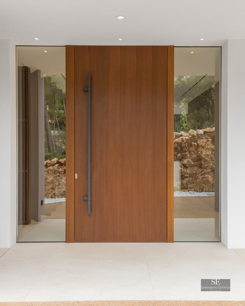 Large wooden pivot door with glass side panels flanked by smooth white walls and minimalist lighting.