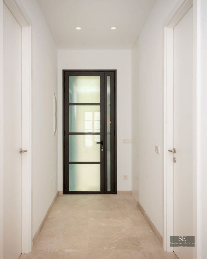 Narrow white hallway with polished stone floors leading to a black-framed glass door.