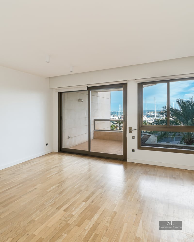 Empty modern room with light wood floors and large windows overlooking a balcony and a marina with palm trees.