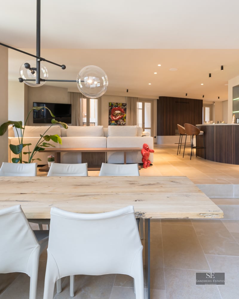 A spacious modern open-plan room featuring a wooden dining table, white chairs, glass globe pendants, and a bar area.