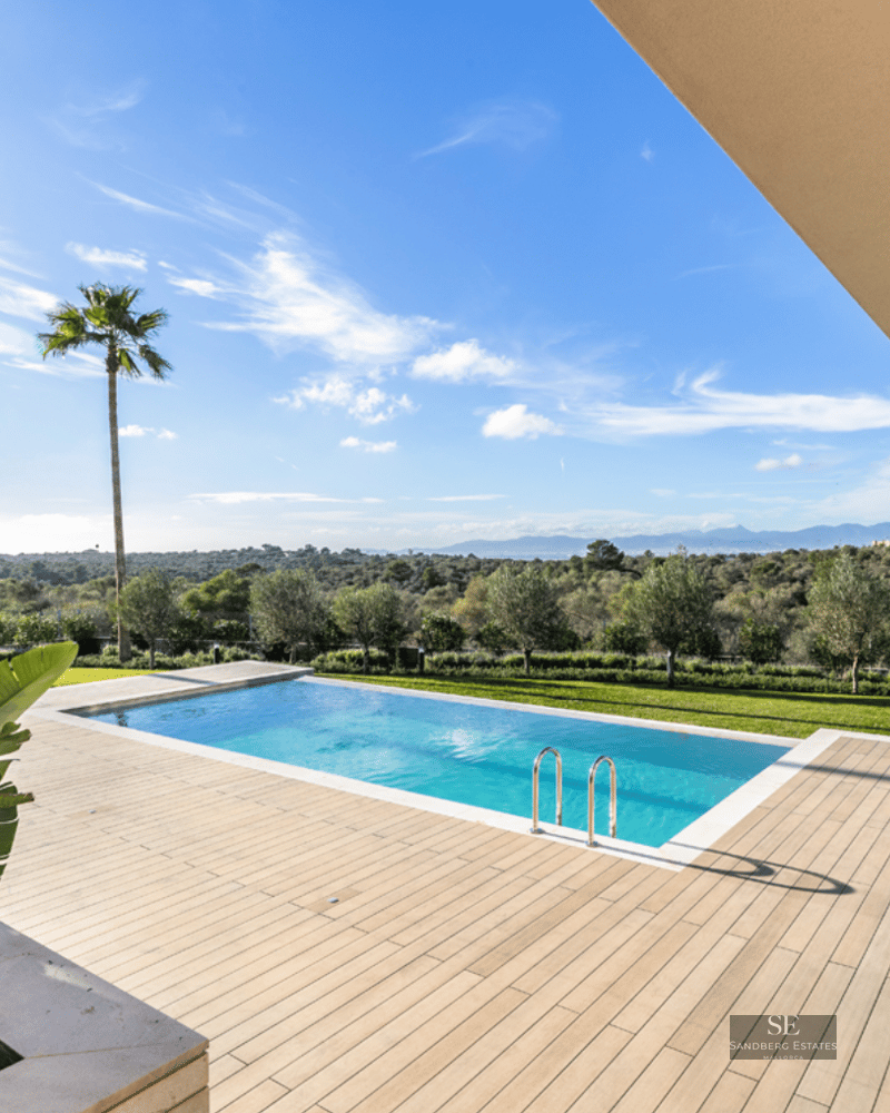 Rectangular blue pool on a light wood deck surrounded by palm trees overlooking a lush green valley.
