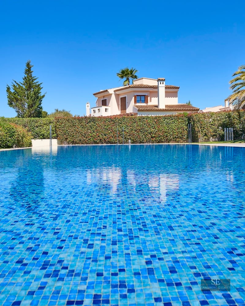 Large blue mosaic pool under a clear sky, with a Mediterranean villa and palm trees in the background.