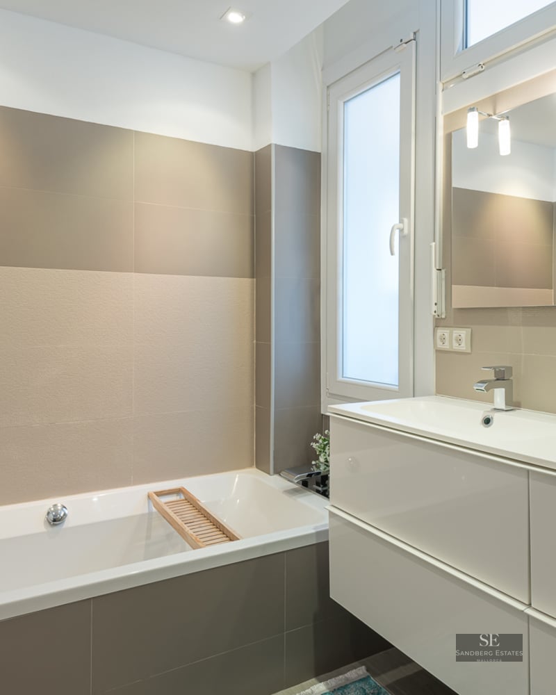 A contemporary bathroom with a white bathtub, a double-sink vanity, large beige tiles, and a wide mirror.