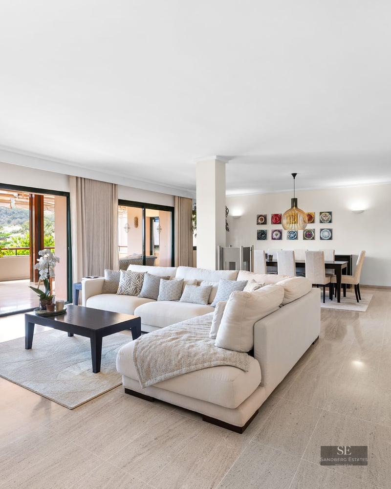 Salon spacieux avec canapé blanc en L, sol en marbre et grandes baies vitrées ouvrant sur une terrasse paysagée.