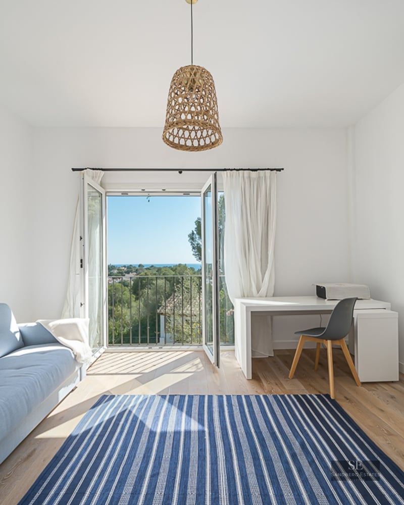 Pièce lumineuse avec canapé bleu, bureau blanc et portes-fenêtres ouvertes sur un paysage verdoyant et la mer.