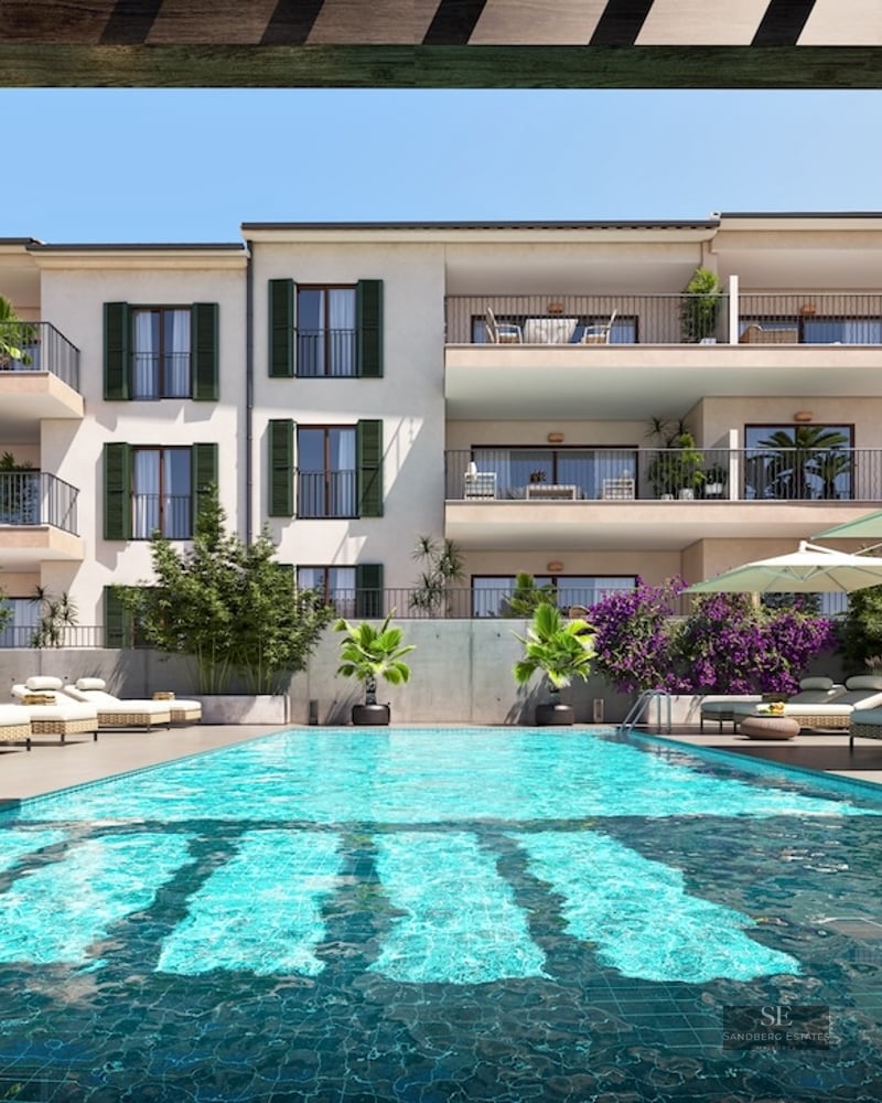 Large turquoise pool with sunbeds and umbrellas in front of a white building with balconies and green shutters.