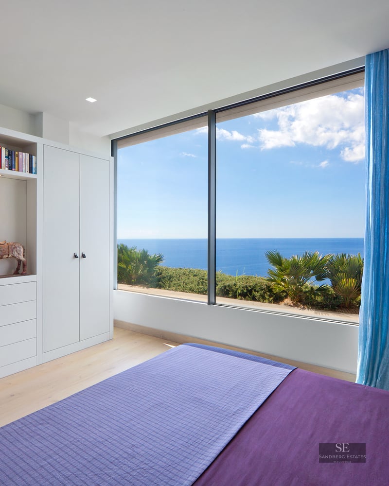 Modern white bedroom featuring a purple bed, large floor-to-ceiling window overlooking the blue sea and sky.