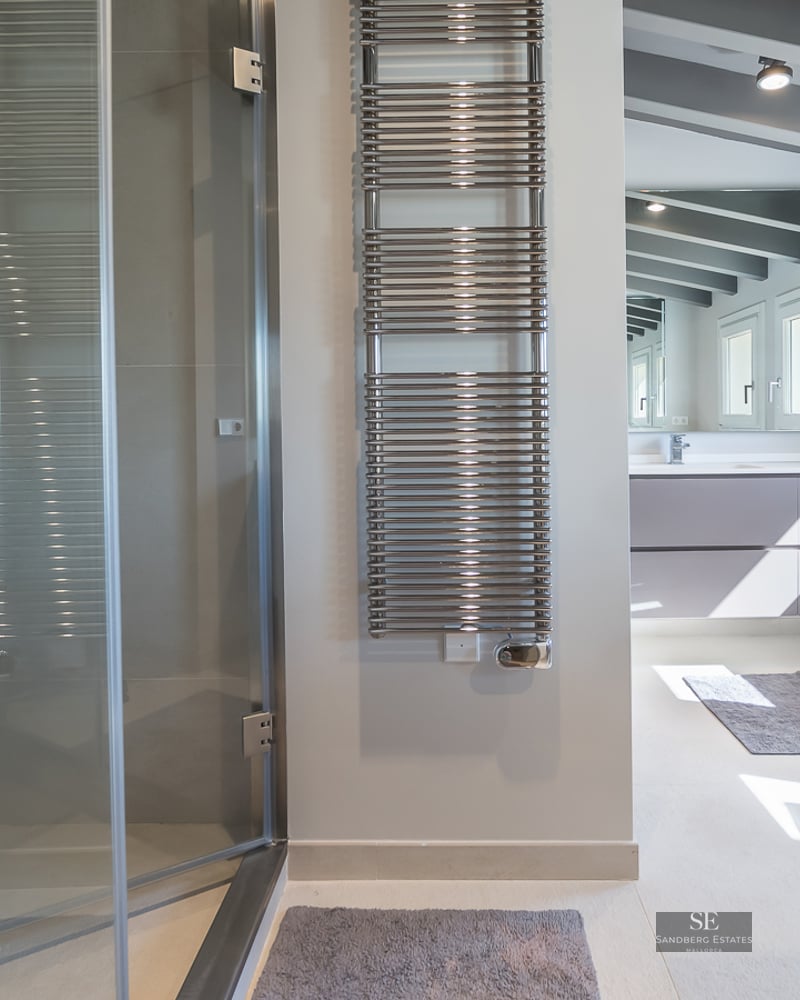 A contemporary bathroom featuring a glass-enclosed shower, chrome towel warmer, and grey ceiling beams.