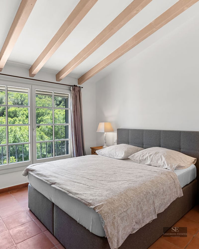 Bedroom featuring a gray double bed, exposed ceiling beams, terracotta flooring, and views of lush green trees.