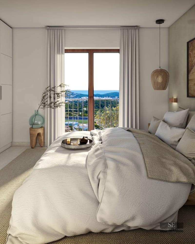 Bright master bedroom with a large bed, built-in white wardrobes, and a window overlooking a coastal landscape.